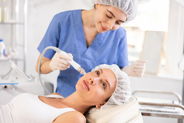 Young female cosmetologist performs cleansing hardware facial procedure on young female patient
