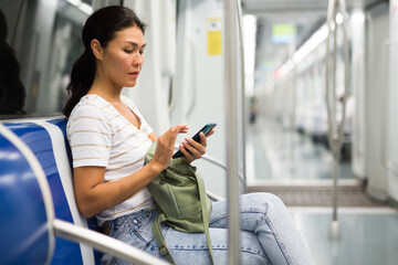 Asian woman with bag and sitting on bench in subway train and using smartphone.
