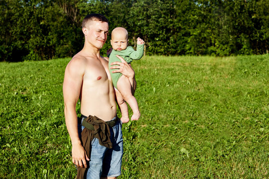 Shirtless European man holds baby in his arms in countryside.
