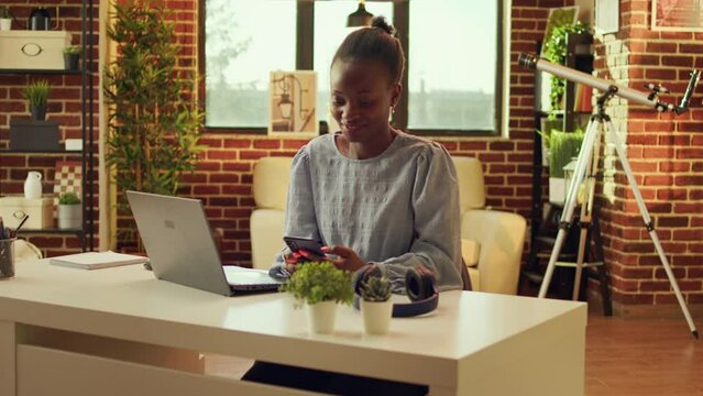 Woman teleworker scrolling webpage and checking social media before starting daily freelance tasks, network connection. African american self employed person sitting at desk at sunset.