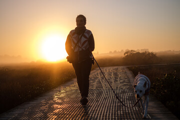 looking into the sun at a woman walking a medium sized dog on a boardwalk through an estuary at dawn in the winter season
