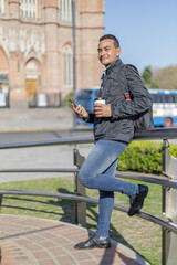 Fototapeta premium Latino man holding a disposable cup of coffee leaning on a railing in a public park.