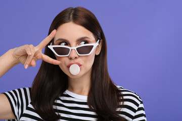 Beautiful woman in sunglasses blowing bubble gum on light purple background, space for text