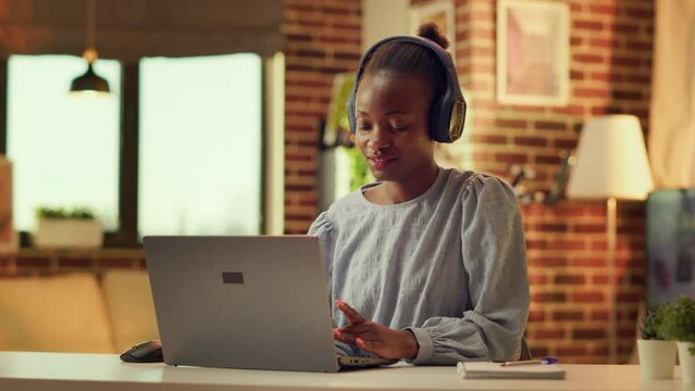 Teleworker listening to music and solving daily tasks, self employed woman. African american girl having fun with headset while she works on duties during sunset hour at home. Handheld shot.