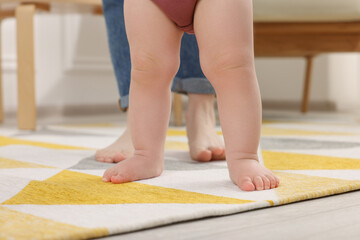 Mother supporting her baby son while he learning to walk on carpet at home, closeup