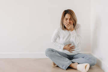Asian Thai woman feeling sick and queasy, has a stomachache cramp, grabbing stomach with one hand and covering mouth in white sweater, sitting on floor in white apartment room in winter alone.