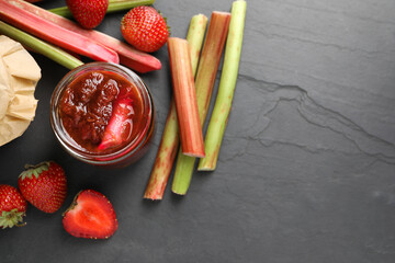 Jar of tasty rhubarb jam, fresh stems and strawberries on dark textured table, flat lay. Space for...