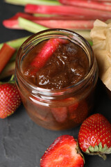 Jar of tasty rhubarb jam, fresh stems and strawberries on dark textured table, closeup