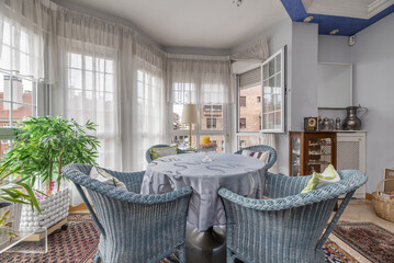 A stretcher table surrounded by blue-painted wicker armchairs next to a large window in an urban...