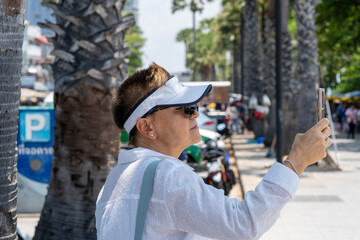An elderly woman in white clothes and sunglasses takes pictures on her phone while standing on a pedestrian sidewalk in the city.