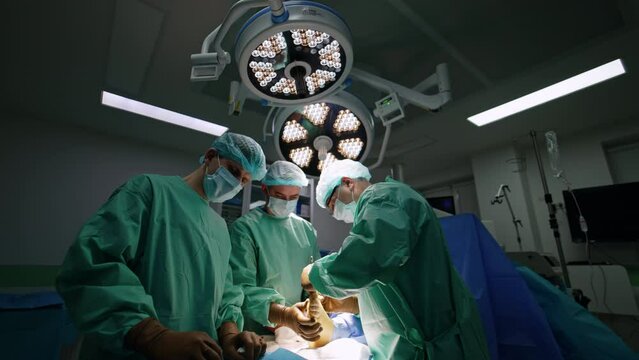 Assisting male doctor holds the foot of a patient helping the main surgeon. Mature surgeon wearing glasses applies metal tools. Male nurse prepares instruments.