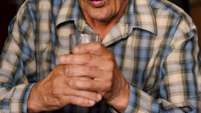 Lonely elderly man drinks water. Hand tremors. Close up hands of senior elderly man with parkinson's disease symptom, hand numbness, finger lock, hand pain. Mental health and elderly care concept.