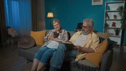Naklejka premium In the picture the elderly couple sit on the sofa in the apartment against the blue wall. The man reads the book aloud. The woman listens, rejoices and drinks tea. Home coziness