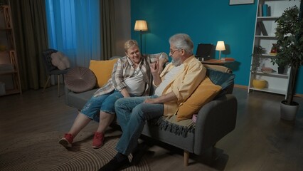 Naklejka premium In the picture, an elderly couple is sitting on a sofa in a room. The man is sitting next to a hidden pink box under a pillow. A gift for a woman, wants to surprise her, she does not see or know