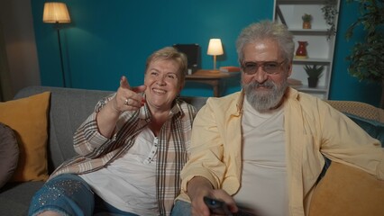 The footage shows an elderly couple sitting on a couch in a room against a blue wall. They are looking away as if at a television. They are happy, laughing