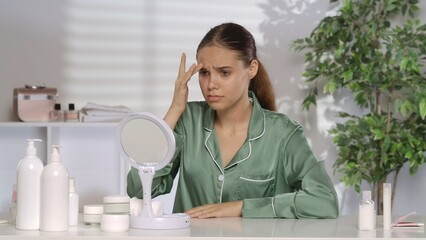 A woman with problematic facial skin looks disappointedly in the mirror. A woman without makeup and wearing green silk pajamas sits at her dressing table in the morning. The concept of problem skin.