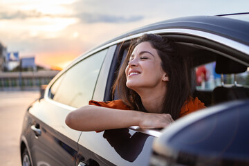 Cheerful young female sitting in shiny car on passenger seat and leaning out open window while enjoying the ride