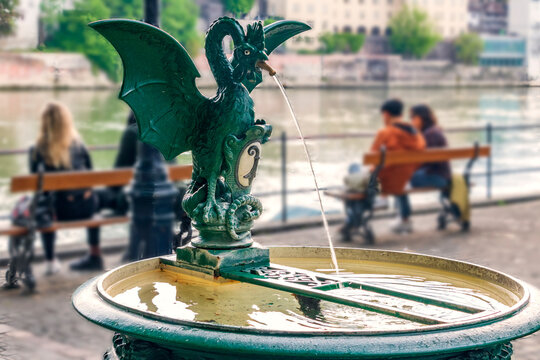Fountain with a sculpture of the Basilisk on the banks of the Rhine in Basel, on the background local people and tourists relax on benches