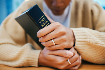 Hands, bible and prayer at desk, religion and Christian worship in home at table. Closeup, holy...