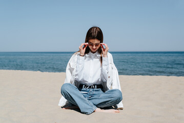 Stylish slender young woman sitting in lotus position on sand at and touching her sunglasses while looking down, casual lifestyle