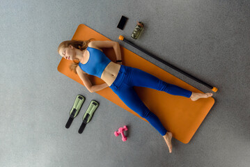 Top view photo of slim sporty woman lying on mat with different sporty equipment