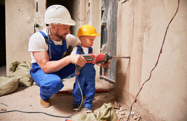 Man and little boy construction workers drilling wall with electric drill in apartment under...