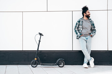 Trendy smiling bearded man in casual clothes riding electric scooter in urban background. Handsome model posing in street near wall. Hipster guy with curly hairstyle. Stands near transport