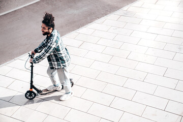 Trendy smiling bearded man in casual clothes riding electric scooter on a road in urban background. Handsome model posing in the street. Hipster guy with curly hairstyle. Top view