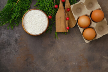 Christmas food background. Ingredients for christmas baking- Flour,eggs, spatula, coniferous branches on dark brown background.