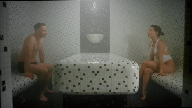 A middle-aged man in boxers and a woman in one-piece swimsuit are in a hammam. They sit in front of each other and have a talk. They are sweating and there is steam in the air.
