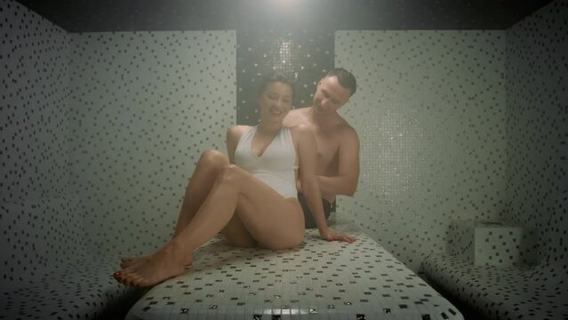 A romantic couple relaxes in a hammam sauna. The woman sits on a tiled bench, she wears a swimsuit. A man is standing behind her. He is massaging her shoulders. She smiles with pleasure.
