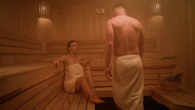 A handsome man in a towel enters a Finnish sauna. The room is wooden. There is a beautiful young woman sitting in the sauna. He sits near her and they look at each other.