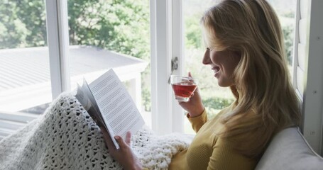 Happy biracial woman sitting next to window drinking tea and reading book at home, slow motion