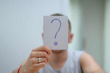 Mężczyzna trzymający na wysokości twarzy kartkę ze znakiem zapytania - obrazy, fototapety, plakaty