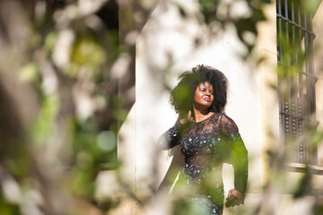 Young woman, beautiful and black with afro hair, with floral dress leaning on a wall, relaxed and quiet, seen through a green plant. Concept beauty, relaxation, tranquility, plants.