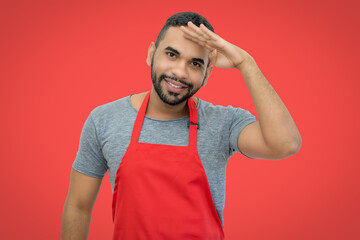 Laughing hispanic waiter with apron on red background looking for guests