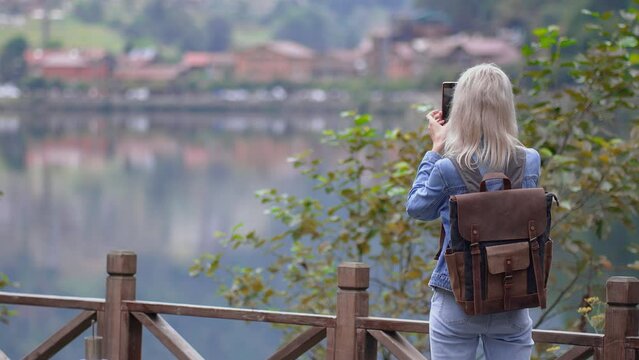 Young beautiful blonde woman in denim suit with backpack traveling alone, taking pictures of beautiful lake Uzungol in Turkey. Concept with thirst for travel, rear view. Blogger Social Media.