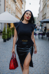 Fashionable dark haired woman dressed in black blouse and leather cut out skirt walking on alley of old city with red purse in hand. Stunning female with red plump lips looking confident outdoors.