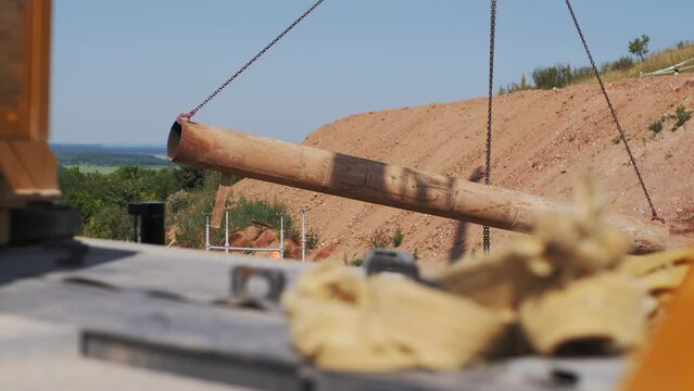 Large tube transported by crane at construction site. Sewer system element hangs on chains near hill. Building material and machinery