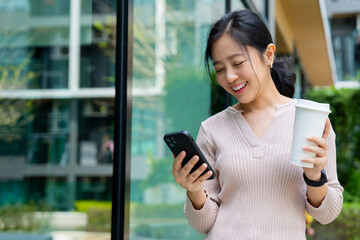 Cheerful Asian female employee stands outside the office using a smartphone and holding a coffee cup.