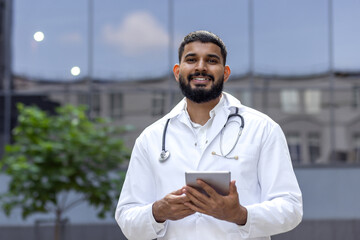 Portrait of a young Muslim male doctor standing in a white coat near a clinic on the street and...