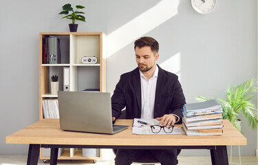 Focused businessman working at home with laptop and papers on desk. Portrait of handsome young man in formal wear sitting at desk in office and looking at laptop screen