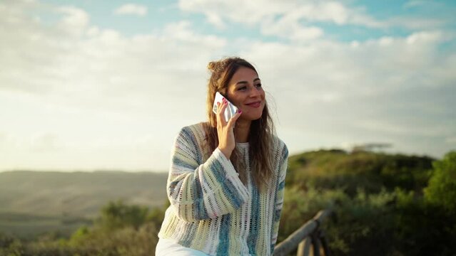Attractive young woman making a cell phone call while smiling with sunset behind her. 