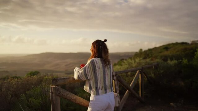 Young brunette model leaning on a wooden railing overlooking hills at sunset.