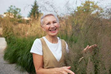 Stylish laughing senior female landscape designer or florist spending time in rural area, walking...