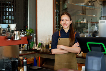 A beautiful Asian business woman in blue shirt stands behind coffee machine at counter. She is smiling and giving a thumbs up. to take pictures to promote a small cafe and boyfriend, business couple.