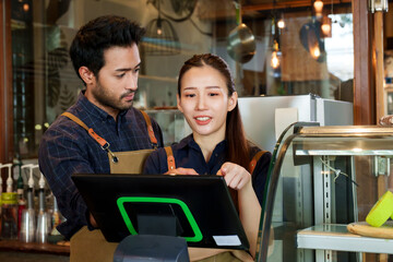 A bearded Indian man wearing overalls teaches how to use a cash register with his beautiful Asian wife. Standing serious behind the cashier counter in coffee shops and bakeries