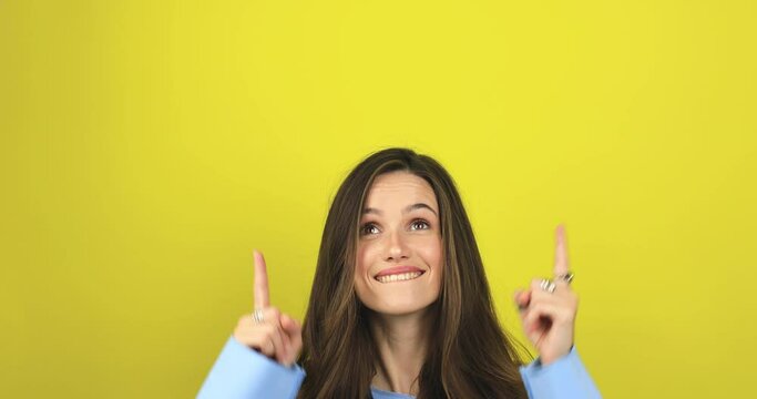 Attractive brunette woman looking at camera and pointing index finger up at copy space for advertising with smile and positive emotions, excited, look up standing isolated yellow background.