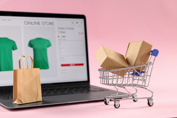 Online store. Laptop, mini shopping cart and purchases on pink background