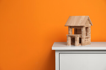 Wooden house on white chest of drawers near orange wall, space for text. Children's toy